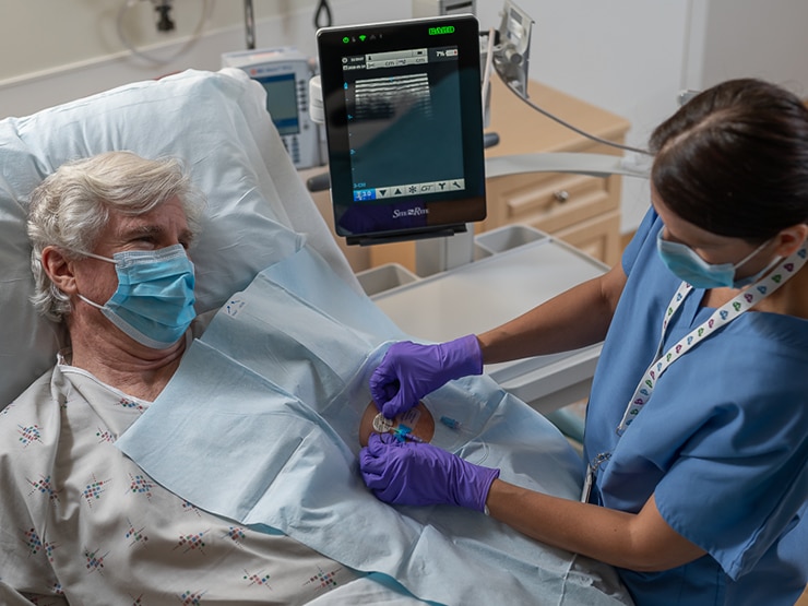 Paciente masculino con mascarilla en una cama de hospital, al que un profesional clínico le administra un dispositivo de acceso vascular