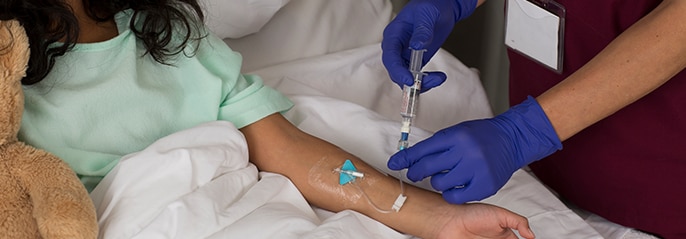niña sentada en una cama de hospital con un osito de peluche, a la que un profesional clínico le administra terapia intravenosa