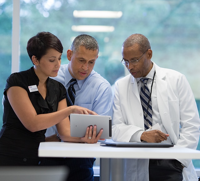 Three healthcare professionals reviewing information on a tablet and paperwork together in a clinical setting.