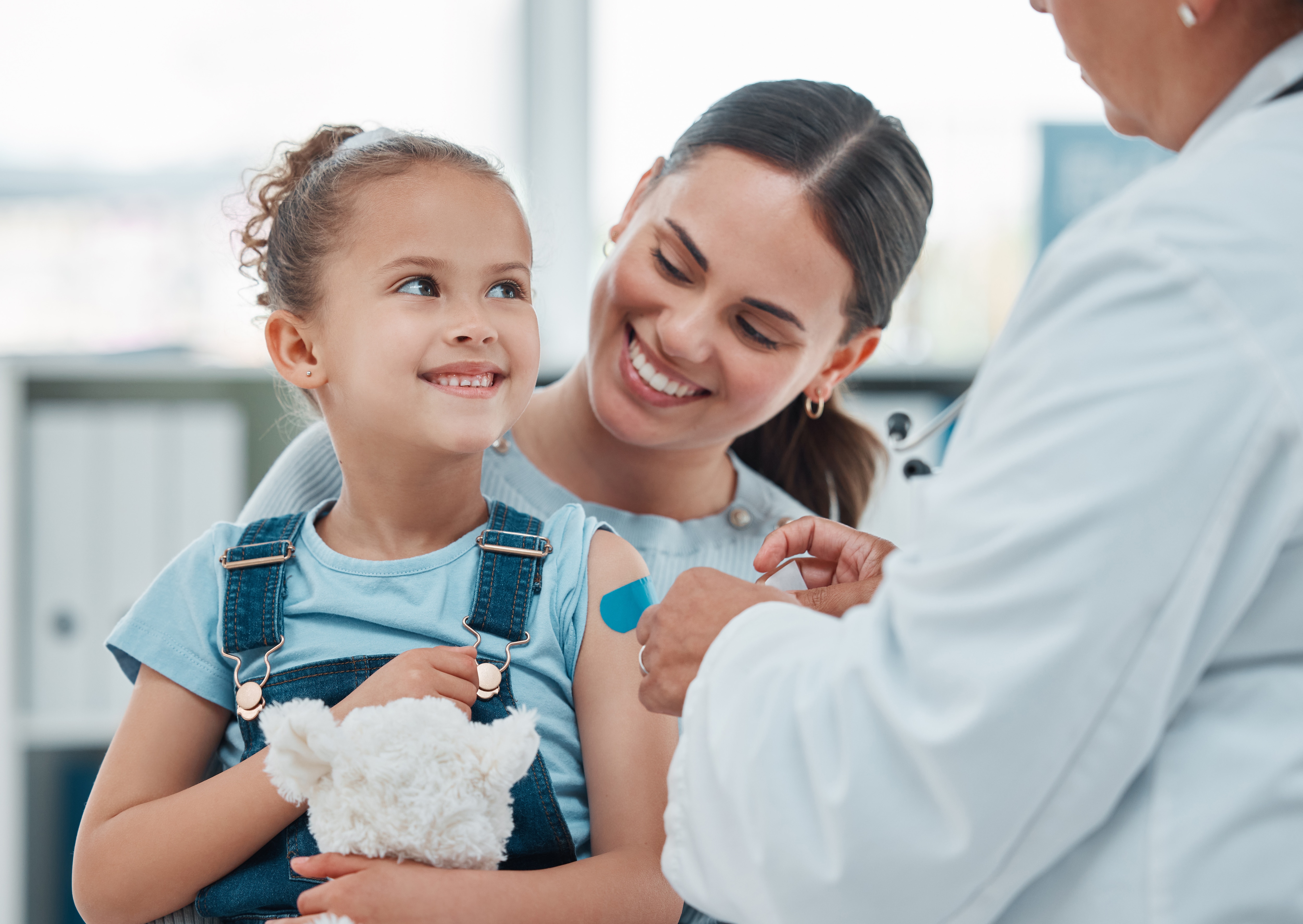 Girl, mom and doctor with vaccine injection, cotton ball and flu shot on arm for disease or covid prevention in hospital. Woman, nurse and child with pediatrician help with bandaid, teddy or health, Girl, mom and doctor with vaccine injection, cotton ball and flu